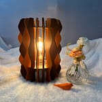 Lampe Pinus en bois à lamelles alternant deux couleurs (Orange et marron). Un oiseau en 3D et un pot de fleurs sont à coté de cette lampe, le tout sur une nappe.