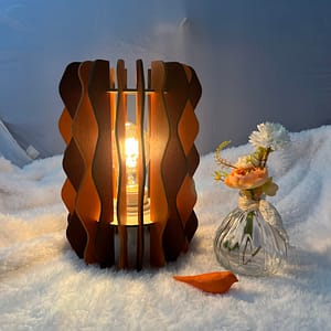 Lampe Pinus en bois à lamelles alternant deux couleurs (Orange et marron). Un oiseau en 3D et un pot de fleurs sont à coté de cette lampe, le tout sur une nappe.