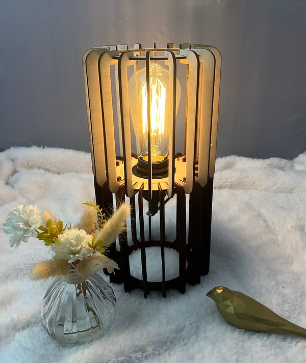 Lampe en bois vanille-chocolat avec son ampoule allumée. Disposée sur une nappe en velour à côté d'un oiseau 3D et d'un vase en verre.