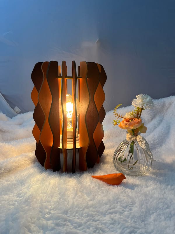 Lampe Pinus en bois à lamelles alternant deux couleurs (Orange et marron). Un oiseau en 3D et un pot de fleurs sont à coté de cette lampe, le tout sur une nappe.