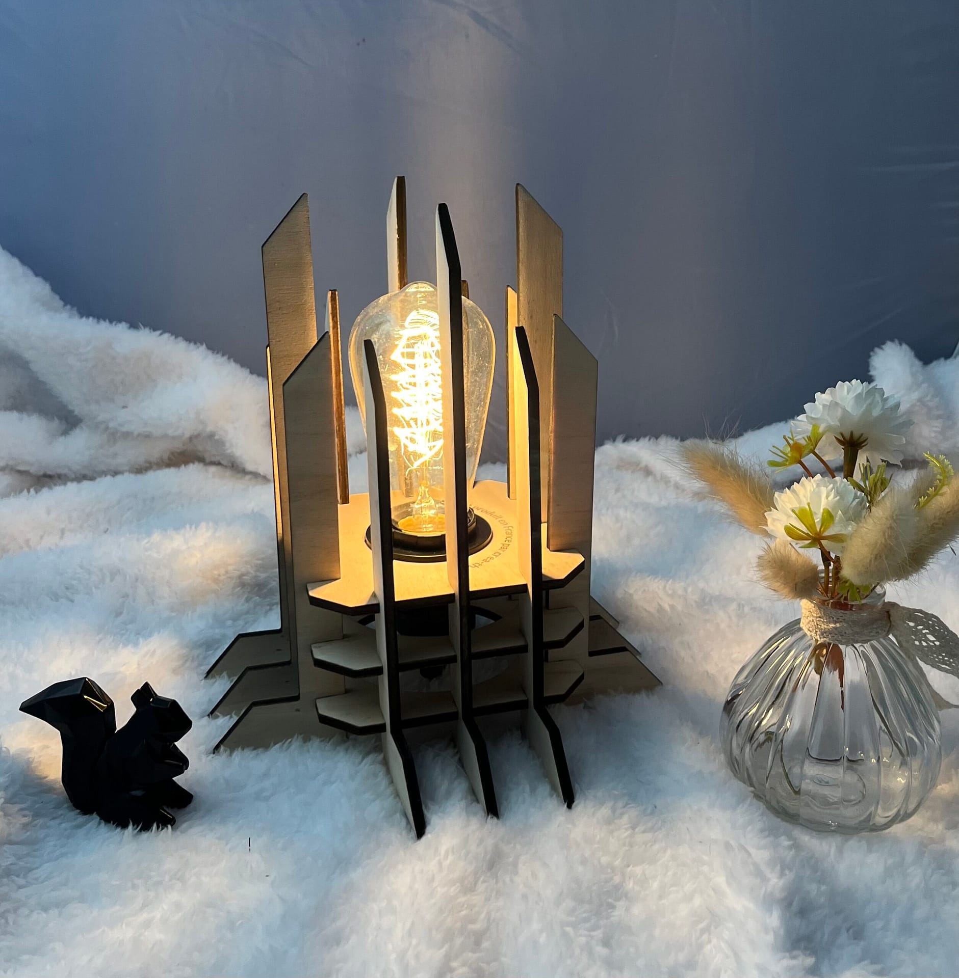 Lampe Luxor en bois moderne. Elle est réversible avec ses deux couleurs noir et blanc. La lampe est posée sur une nappe en velour avev un écureuil en 3D et un pot de fleur à côté