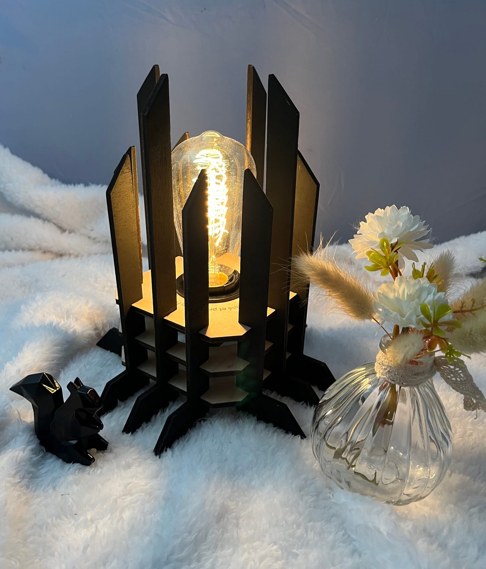 Lampe Luxor en bois moderne. Elle est réversible avec ses deux couleurs noir et blanc. La lampe est posée sur une nappe en velour avev un écureuil en 3D et un pot de fleur à côté